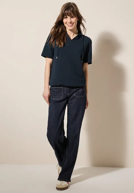 Frau mit dunklem Shirt und Jeans steht lächelnd vor hellem Hintergrund.